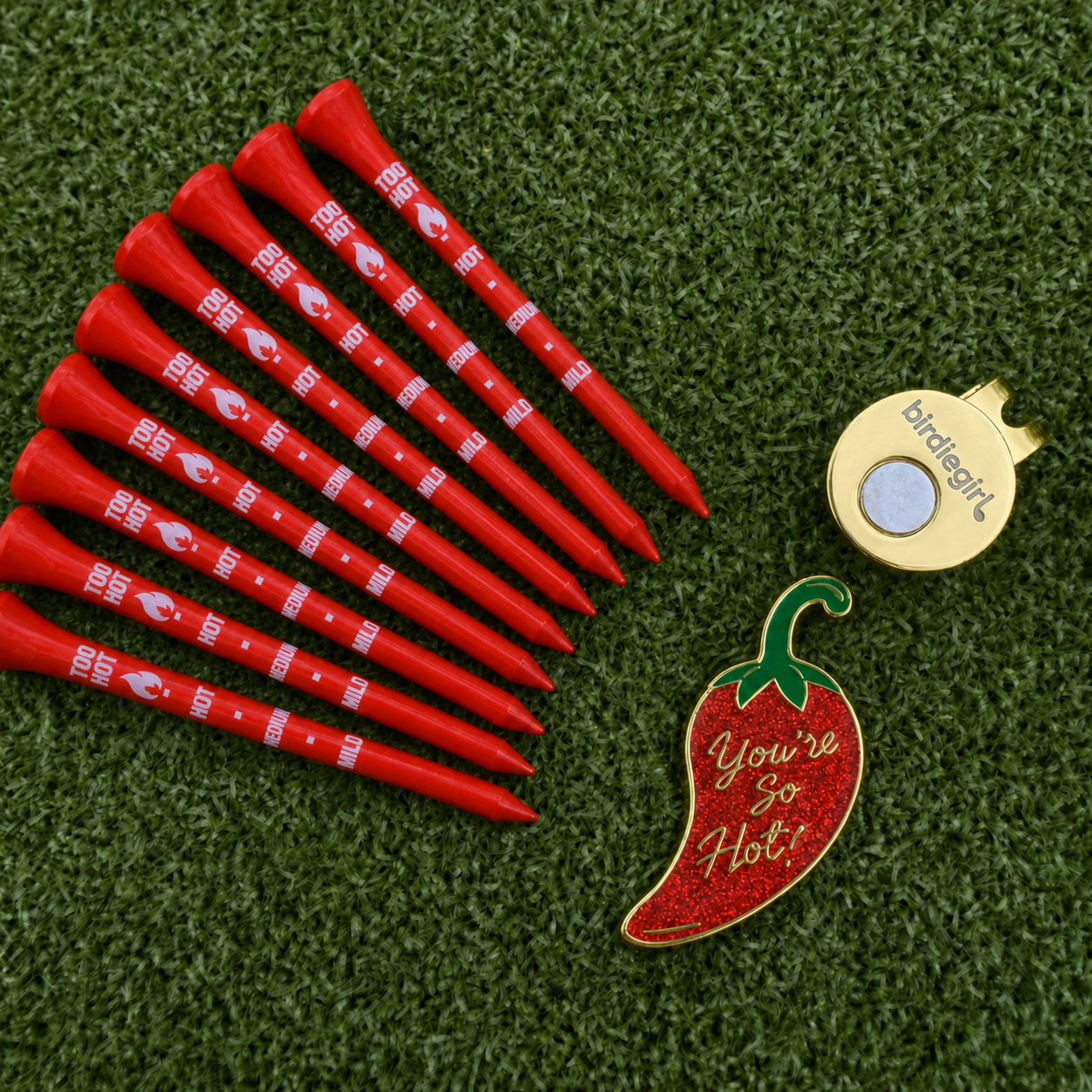 A set of red golf tees with white printed branding, a gold birdie tag, and a red chili pepper-shaped pin reading 'You're So Hot!' arranged on a green artificial turf background.