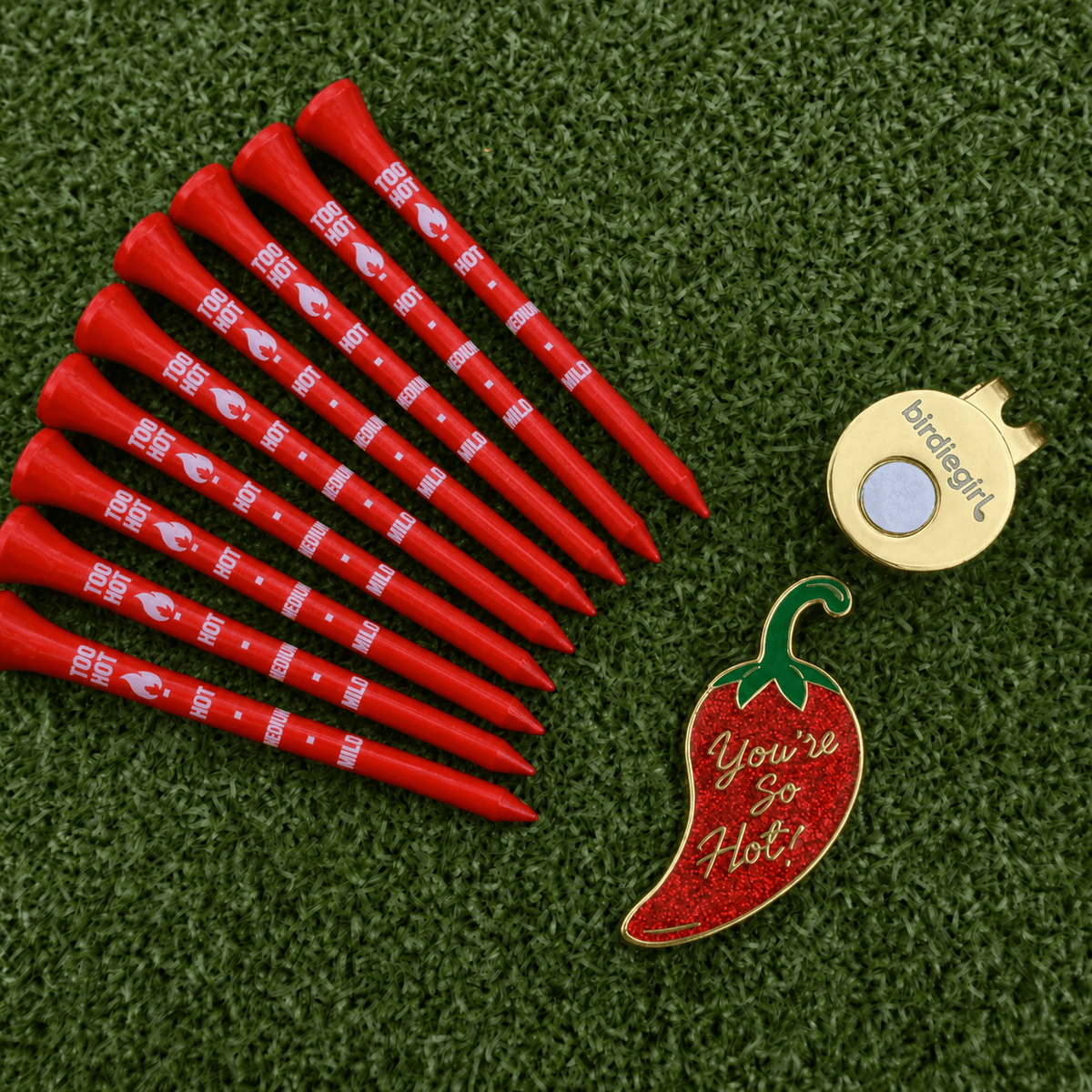 A set of red golf tees with white printed branding, a gold birdie tag, and a red chili pepper-shaped pin reading 'You're So Hot!' arranged on a green artificial turf background.
