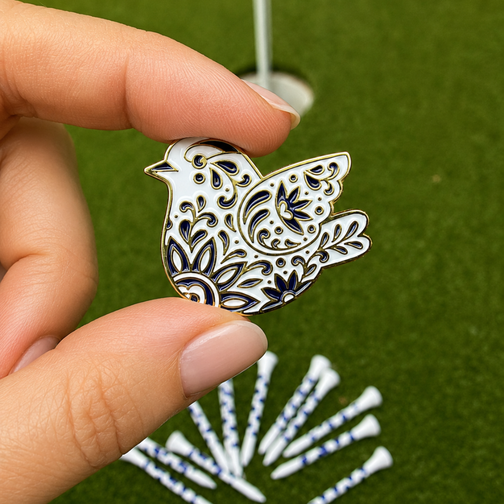 A hand holds a decorative white and navy bird-shaped golf ball marker with intricate cutout patterns, set against a green turf background with golf tees and a flagstick blurred in the distance.