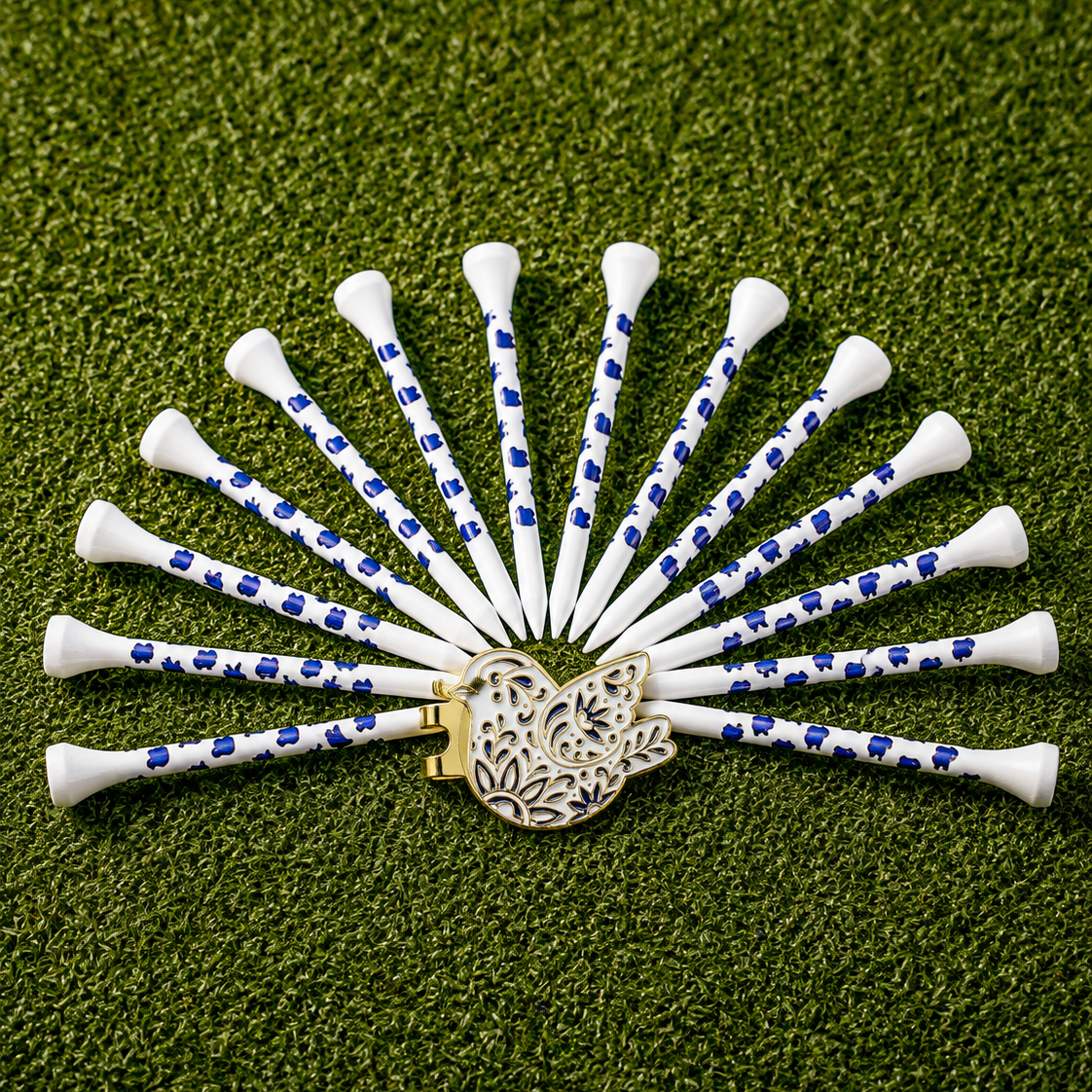 A set of golf tees arranged in a fan shape on artificial green grass, featuring white tees with blue patterns and a decorative gold-toned golf ball marker